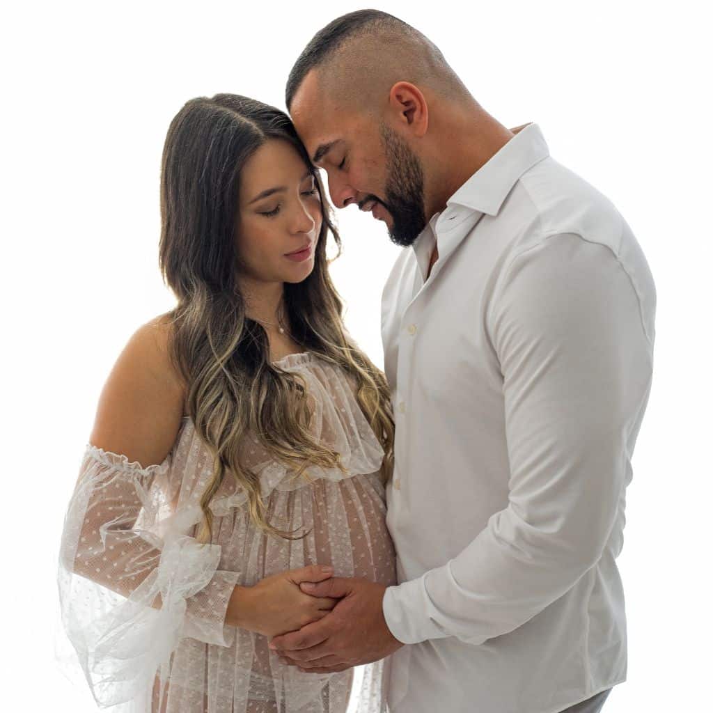 Futuros padres abrazados durante su sesión de maternidad en el estudio, vistiendo ropa blanca y sencilla para un acabado atemporal.