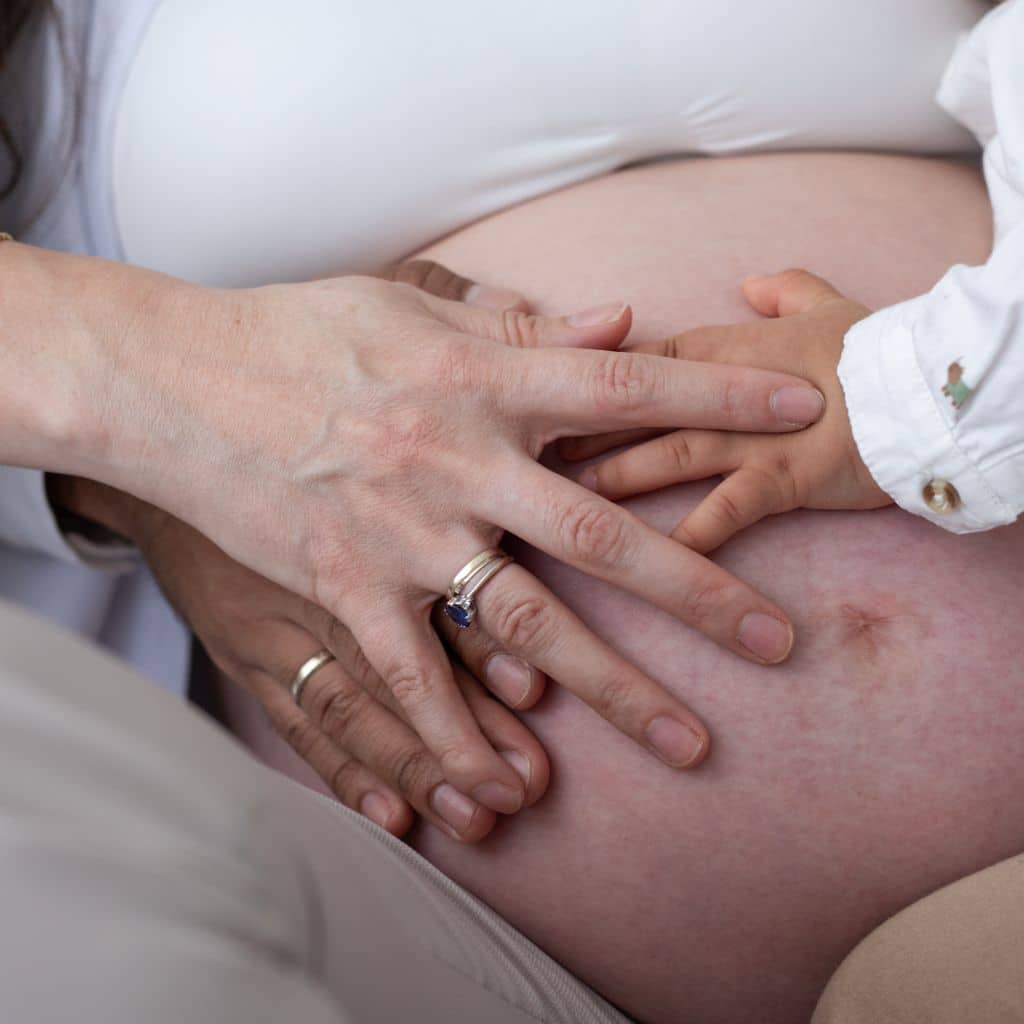 Primer plano de manos de una pareja y un niño pequeño apoyadas sobre la barriga de una mujer embarazada en una sesión de fotos familiar